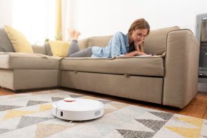 Aspirateur robot laveur blanc en fonctionnement sur un tapis dans un salon, pendant qu’une femme lit sur un canapé